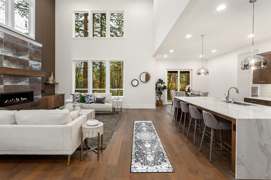 A combined living room and kitchen has 10 windows, wood and marble accents, white walls, couch and a runner rug that divides the rooms.